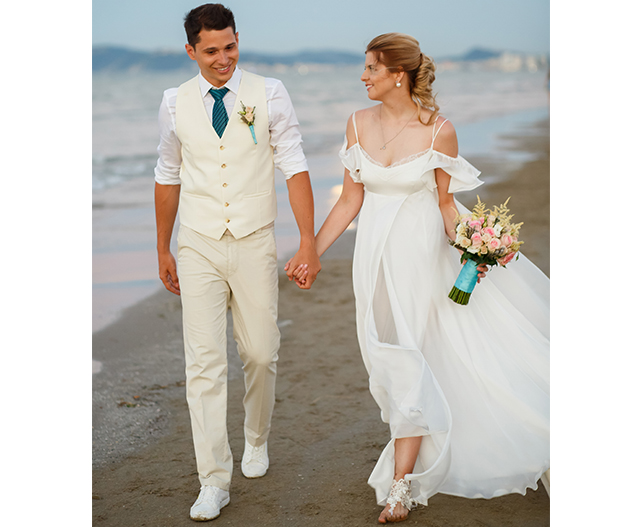 ベスト姿の男性とエンパイアラインのドレスを着た女性がビーチで手を繋いで歩く様子