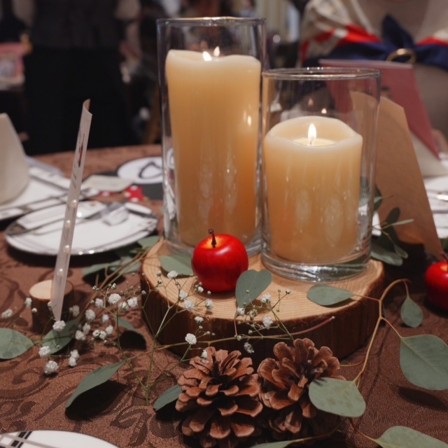 切り株状の木材の上にキャンドル、小さいリンゴの置物が飾られており、そのまわりにはグリーンと松ぼっくりなどが見える