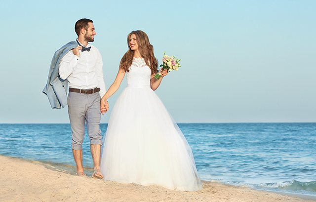 男性とボリュームラインのドレスを着た女性がビーチで手を繋いで歩く様子